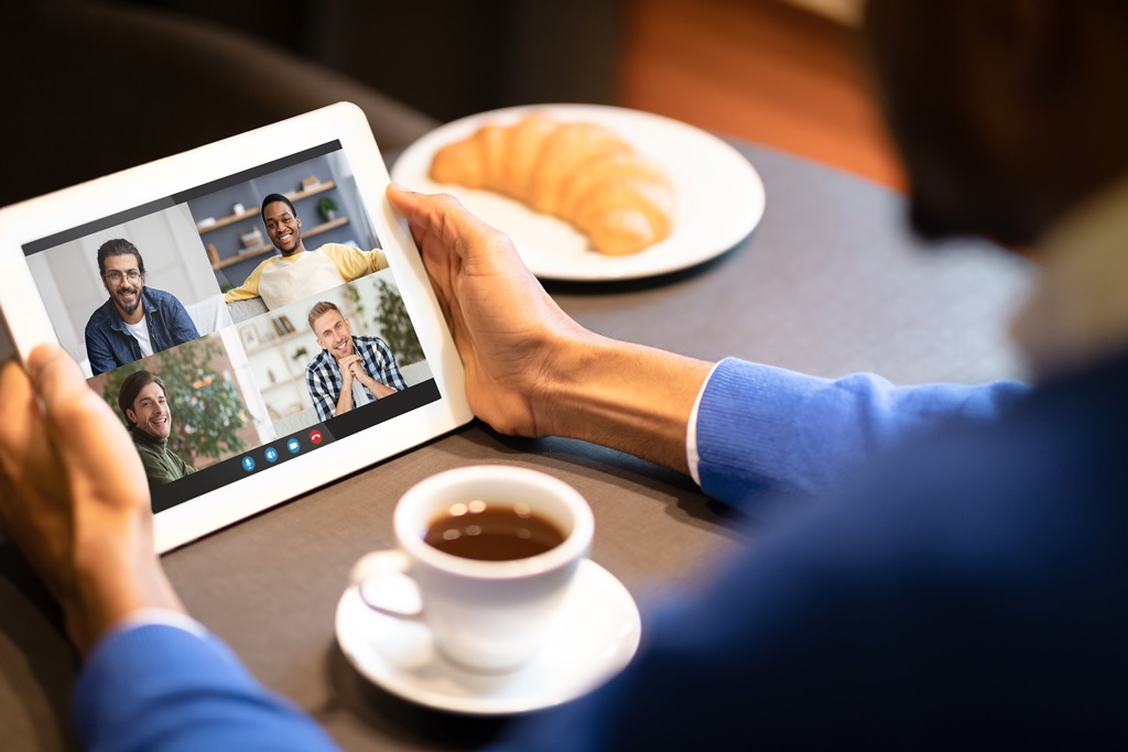 Eine Person sitzt mit einem Kaffee am Tisch und hält ein Tablet während eines online Meetings in den Händen